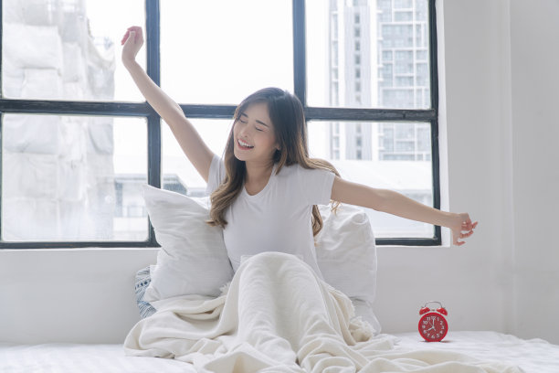 健康的亚洲女性清晨醒来在窗边放松肌肉。快乐小女孩伸展举起手臂肌肉身体舒适，醒来后坐在床上用枕头，闹钟在公寓。图片下载