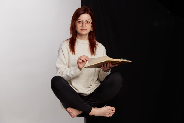 美丽的红发年轻女子读书，而坐在椅子上，双腿交叉超过一半的黑白背景图片下载