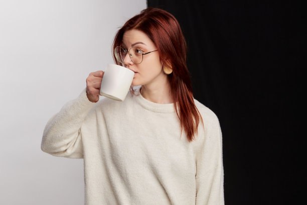 热饮使人精神焕发。红头发的女人手里拿着一杯茶或咖啡，表情站在半白半黑的背景上，戴着大眼镜图片下载