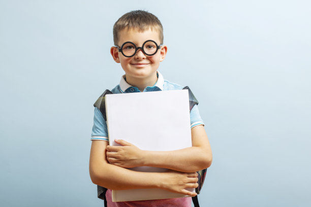 回到学校。快乐的小学生戴着眼镜拥抱着一本书，对着蓝色背景的相机微笑。自由空间图片下载