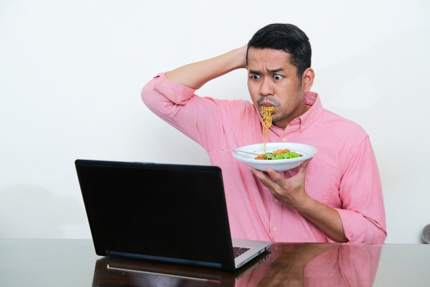 一名亚洲成年男子一边吃面条，一边用震惊的表情看着自己的笔记本电脑图片下载