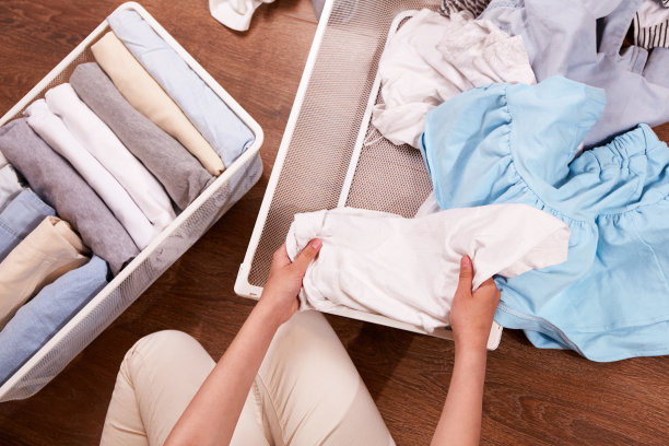 一个女人整理一堆衣服的手。附近地板上的金属筐里堆着一堆堆衣服。图片下载