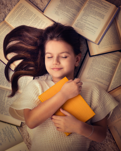 女孩躺在地板上，抱着一本黄色的书。她的脑袋周围都是书。女孩在做梦。她读了很多书。图片下载