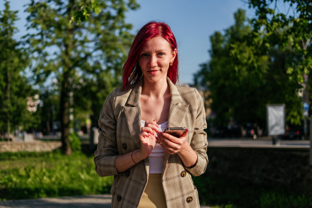 红色头发微笑的女人手持智能手机站在户外。女孩用智能手机看相机图片下载