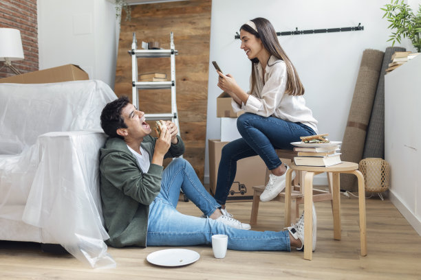 一对可爱的情侣在他们的新房子里，围着纸板箱一边吃着三明治，一边用智能手机拍着有趣的照片图片下载