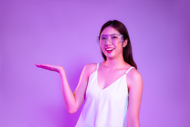 年轻的亚洲女性穿着白色背心，戴着眼镜，在紫色的屏幕背景上摆出展示和展示的姿势。图片下载
