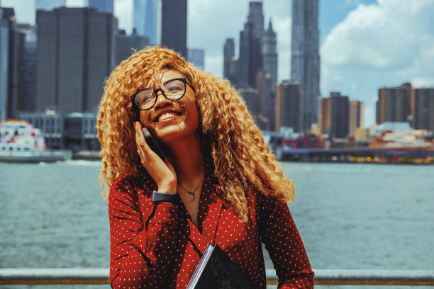 照片中，年轻的成年企业家千禧一代女性戴着眼镜，留着爆炸头，微笑着在户外打电话，身后是纽约曼哈顿的天际线图片下载