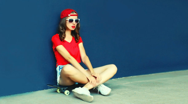 现代年轻女子坐在滑板上戴着棒球帽，短裤在城市街道上空白拷贝空间蓝色的墙背景图片下载