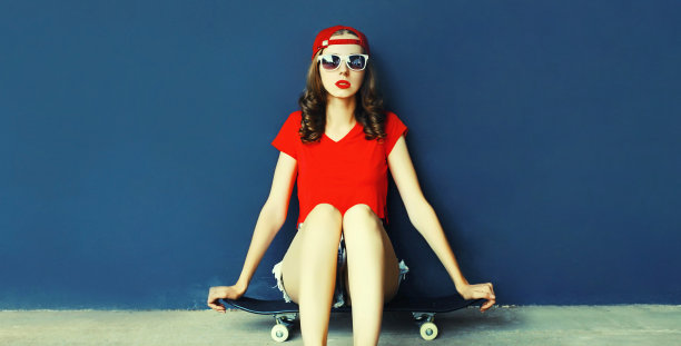 现代年轻女子坐在滑板上戴着棒球帽，短裤在城市街道上空白拷贝空间蓝色的墙背景图片下载