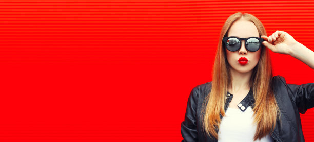 肖像的金发年轻女子吹她的嘴唇与口红发送空气吻戴墨镜在红色的背景，空白的拷贝空间的广告文本图片下载