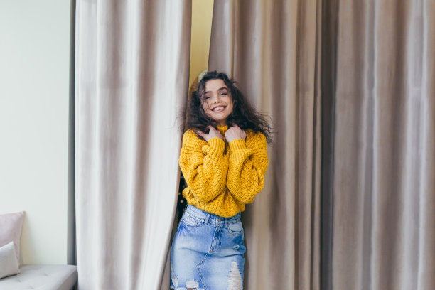 一个年轻漂亮的女孩穿着新衣服，穿着黄色温暖的毛衣和牛仔裙在一家服装店的肖像，背景是试衣间附近的棕色窗帘，摆姿势，展示，炫耀，看着镜头，微笑图片下载