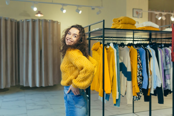 一个年轻美丽的卷发女人的肖像，摆姿势，穿着和摆姿势的新衣服在黄色温暖的毛衣和牛仔裙在一家服装店图片下载