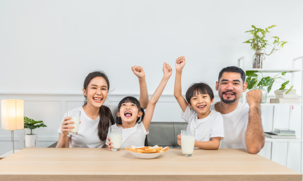 亚洲家庭在客厅享用早餐。小女孩女儿坐在桌子上，和微笑的爸爸妈妈在早上喝牛奶。家庭幸福。图片下载