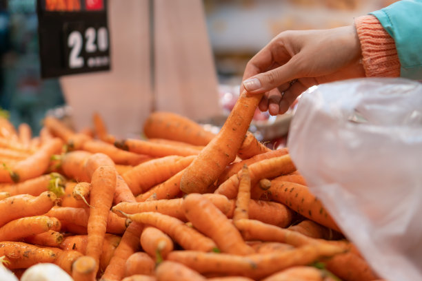 女人的手在商店里用玻璃纸包着新鲜胡萝卜的特写。美味健康的食物图片下载