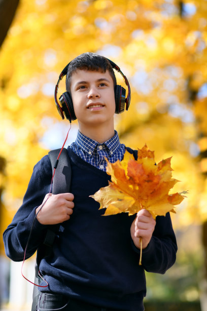 一个十几岁的男孩走在秋天的公园里，戴着耳机听着音乐，阳光明媚的日子里，树上的叶子都泛黄了图片下载