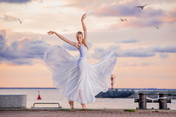身穿白色飞行裙和紧身裤的芭蕾舞演员，在海堤或海滩上以夕阳的天空为背景图片下载