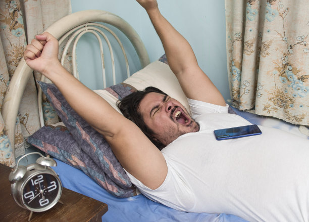 一个睡眼惺忪的人躺在床上打哈欠，伸开双臂。被闹钟吵醒了。早上的场景。图片下载