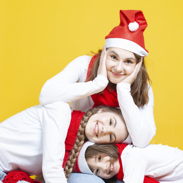 季节性节日的想法。三个可爱的微笑快乐的高加索家庭女孩在圣诞老人的帽子有乐趣，而拥抱对方在时尚的黄色背景。图片下载