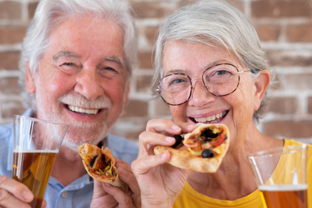 老夫妇在家里一起吃披萨，看着镜头，砖墙背景图片下载