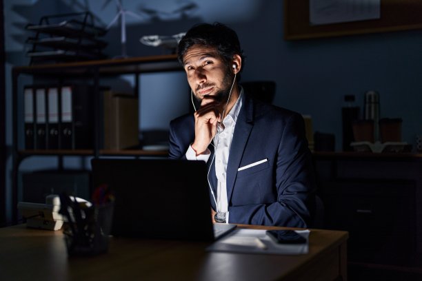 英俊的拉丁人在办公室工作在晚上严肃的脸思考问题与手放在下巴，深思困惑的想法图片下载