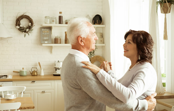 快乐迷人的老妇人微笑着对着镜头，与她的丈夫在厨房里跳华尔兹。图片下载