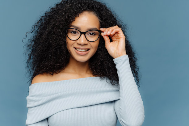 讨人喜欢的年轻非洲裔美国女人的头像保持手在框架眼镜，有温柔的微笑，穿蓝色套头衫，有最小的妆容，健康的皮肤，隔离在蓝色的背景图片下载