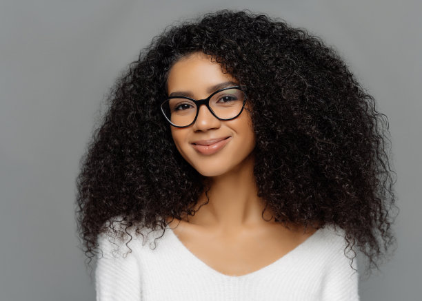 特写肖像的高兴美丽的非洲妇女与浓密的卷发，通过透明的眼镜，穿白色毛衣，对工作室的灰色墙壁的姿势。种族，美貌，面部表情图片下载
