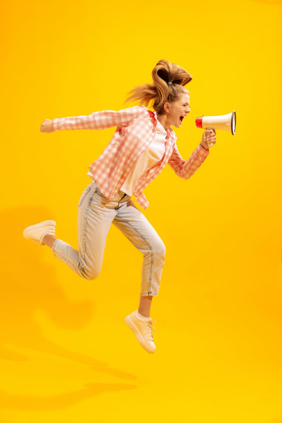 年轻的情绪化的女孩的肖像摆姿势，大喊大叫的扩音器孤立在黄色的工作室背景。新闻图片下载