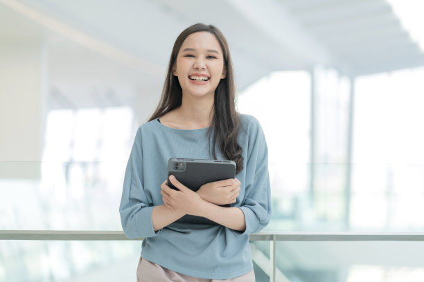 亚洲微笑开朗女性数码游牧民手持平板设备看相机人像拍摄，幸福微笑的亚洲女性站在办公室学院走廊积极微笑的态度图片下载