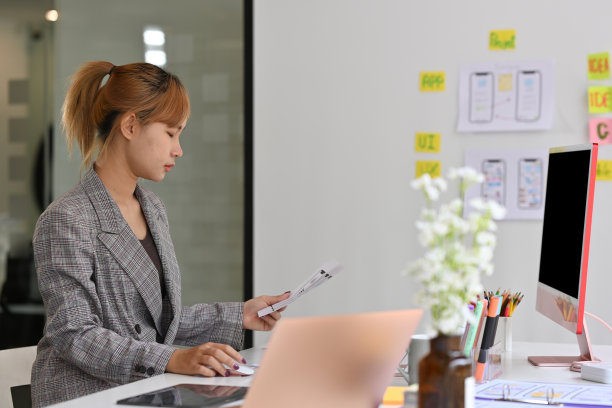 泰国亚洲女性在电脑上工作的侧视图肖像，在现代办公室内部的布局纸上看起来若有所思。技术和设计理念。图片下载