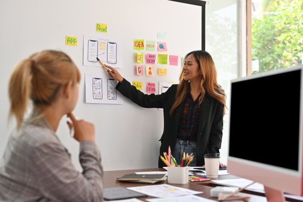 这位亚洲女商人指了指白板上一个媒体设计的布局，这个布局是为了在黑屏电脑旁与另一位同事分享想法。技术和设计。图片下载
