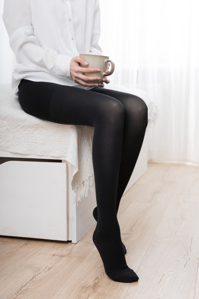 美丽的年轻女子坐在床上压缩丝袜与一杯咖啡或茶在她的手，特写。穿着黑色丝袜的女性长腿。女孩穿上长袜。黑色紧身裤图片下载