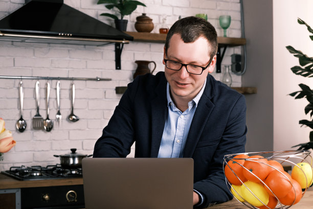 一个年轻的成功商人的肖像使用笔记本电脑坐在家里的厨房里，穿着商务西装，戴着眼镜。一名男子微笑着看着水果篮旁的笔记本电脑显示器图片下载