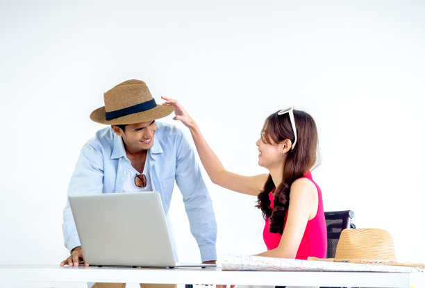 幸福的亚洲夫妇，年轻的女人把帽子戴在男人的头上微笑着，同时用笔记本电脑在桌子上一起为旅行信息在家里办公，准备旅行，快乐的暑假概念。图片下载