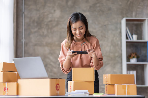 年轻的亚洲女性用手机或智能手机数码相机拍下邮箱地址的照片，在网上出售，并准备包装产品包装盒。销售中小企业网上想法概念图片下载