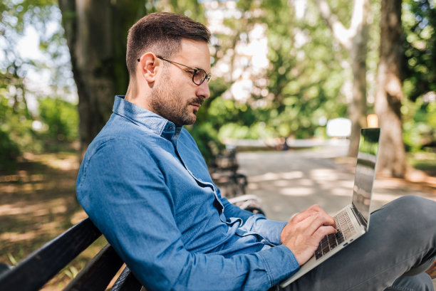 严肃的企业家坐在公园的长椅上用笔记本电脑工作。图片下载