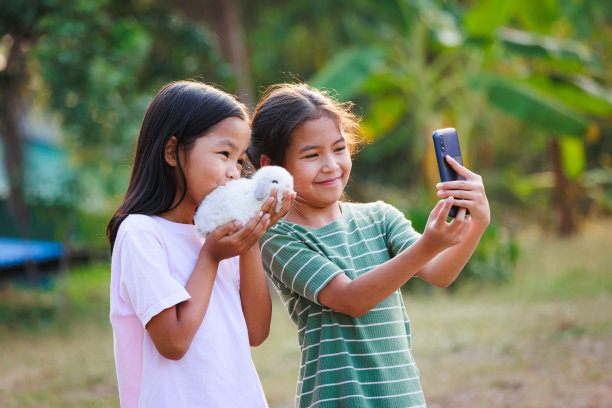 两个亚洲女孩拍一张照片和玩可爱的兔子。姐妹俩手里拿着一只小兔子霍兰德，一起在花园里用智能手机自拍，玩得很开心。图片下载