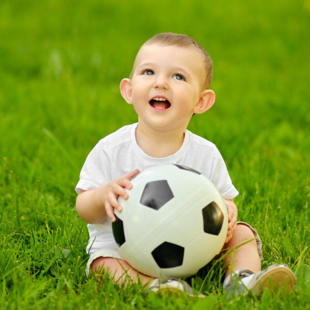 快乐的蹒跚学步的小男孩正在绿色的草地上玩足球。穿着白色衣服的微笑的孩子手里拿着一个球坐着，一岁大图片下载