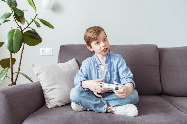 穿着蓝色衬衫的快乐男孩坐在沙发上，手里拿着白色的操纵杆玩电子游戏。在家玩电子游戏的概念图片下载