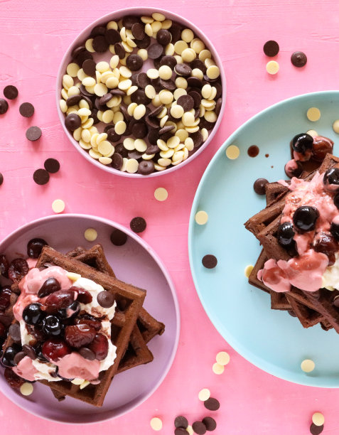 特写图片:自制的巧克力华夫饼堆叠在一起，上面放着融化的覆盆子冰淇淋、蓝莓和樱桃，碗状牛奶和白巧克力片，淡紫色和浅蓝色的盘子，粉红色的背景，俯瞰风景图片下载