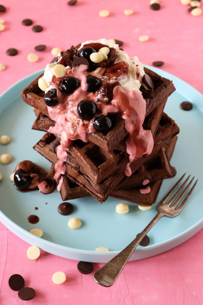 特写图片:一堆自制的巧克力华夫饼，上面放着融化的覆盆子冰淇淋、蓝莓和樱桃、牛奶和白巧克力片，淡蓝色的盘子、叉子、粉红色的背景，升高的视野，聚焦前景图片下载