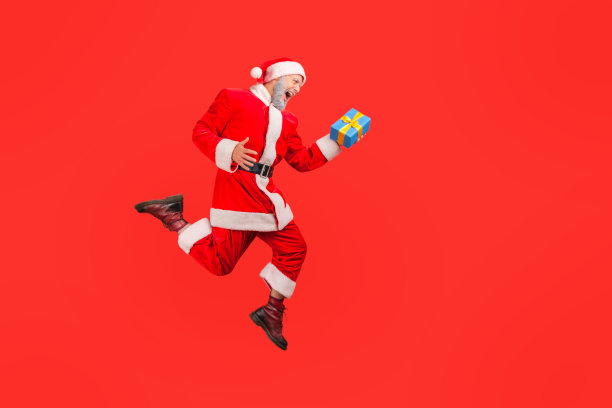侧视图全长老人与灰色胡子穿着圣诞老人服装跳跃在空中与礼物盒，庆祝圣诞节。图片下载