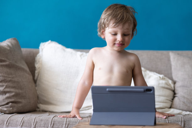 可爱的金发蹒跚学步的孩子看动画片或玩游戏用平板电脑，坐在家里的沙发上。孩子们的小玩意和现代娱乐。视频聊天，视频通话。图片下载