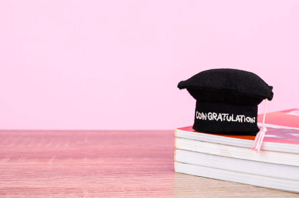 教育理念:毕业帽或学位帽放在许多书和放置在木制桌子孤立的粉红色粉彩背景。图片下载