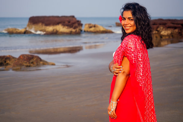 快乐和美丽的印度女人在传统时尚的红色纱丽手持相机为摄影师在海滩热带天堂海洋摆姿势图片下载