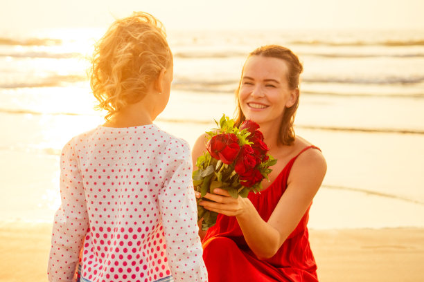金发小女孩给了一束玫瑰在海滩上惊喜和惊奇的母亲。美丽的母女带着鲜花在海边。母亲节快乐。情人节2月14日图片下载
