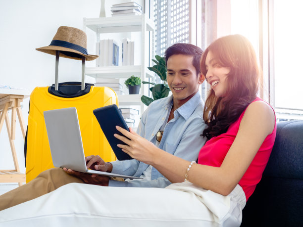 幸福的亚洲夫妇，年轻的男女看着和使用笔记本电脑和智能手机一起预订机票和旅行信息在客厅附近的行李，旅游假期暑假。图片下载