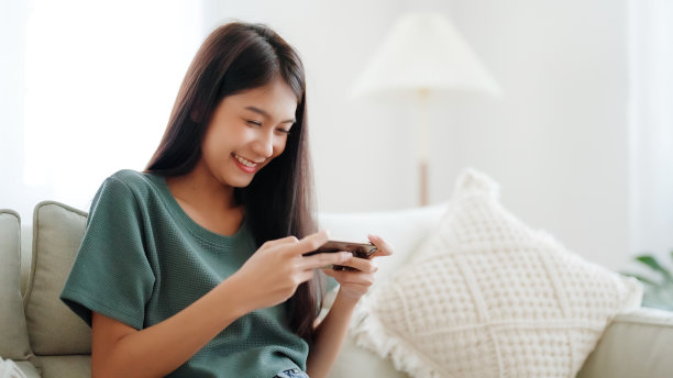 快乐的年轻亚洲女性放松在舒适的沙发在家里的智能手机短信，微笑的女孩使用手机聊天，浏览无线网络小工具，从家里在线购物图片下载