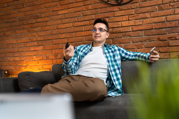 一名年轻的白人男性坐在家里的沙发沙发床上用遥控器看电视拷贝空间图片下载