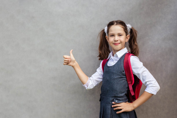 一个十几岁的女孩的特写肖像在学校的制服和背包背后，她的一个手指在灰色的背景上图片下载